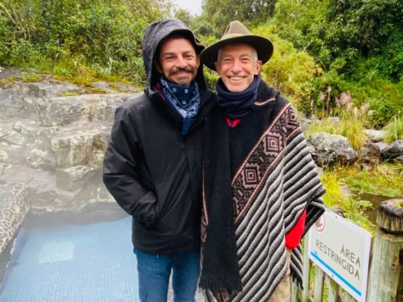 Ben Hoffman and his godfather smiling together on a misty outdoor trail in South America, one in a rain jacket and blue bandana, the other in an Andean poncho and wide-brimmed hat