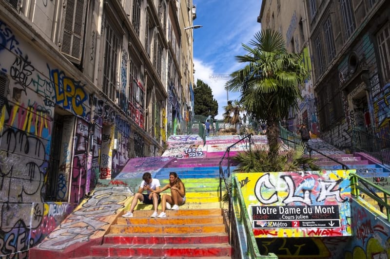 Rainbow-painted staircase and graffiti-covered walls in the Cours Julien neighborhood of Marseille, France, with two people sitting on the colorful steps