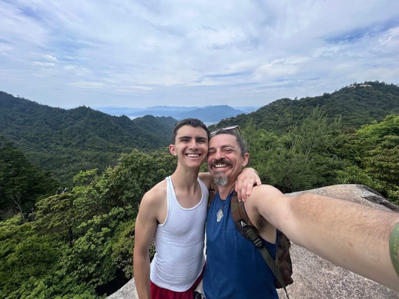 Ben Hoffman and a young companion smiling at a rocky mountain viewpoint in Japan, forested hills and distant valley visible behind them