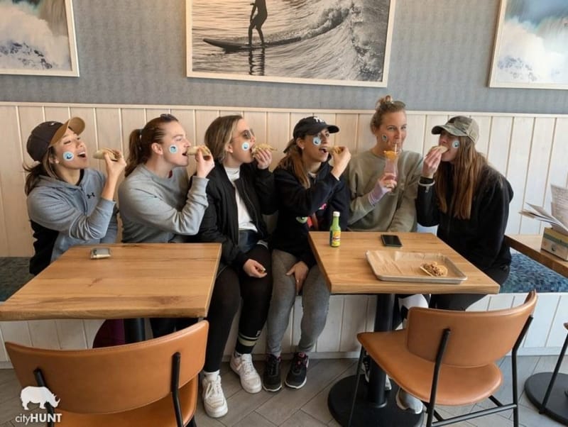 Six women eating tacos at a coastal restaurant, each wearing a cityHUNT sticker on their face, at a Long Beach Chamber of Commerce team-building event